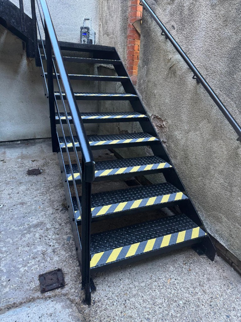 Lower flight of the external steel staircase painted black with anti-slip nosings on a clean floor — after