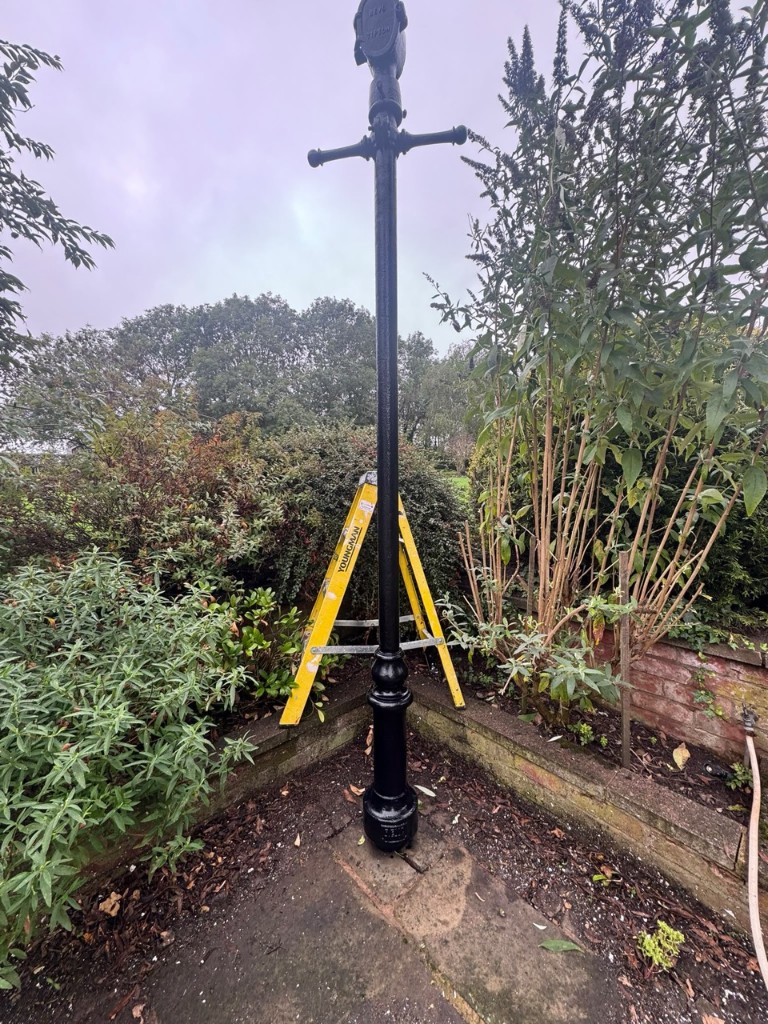 Tall cast iron lamppost freshly painted gloss black in a garden corner with stepladder nearby