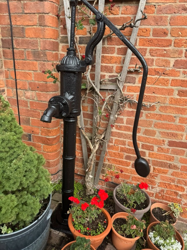 Freshly painted black cast iron water pump against a brick wall with garden pots at the base