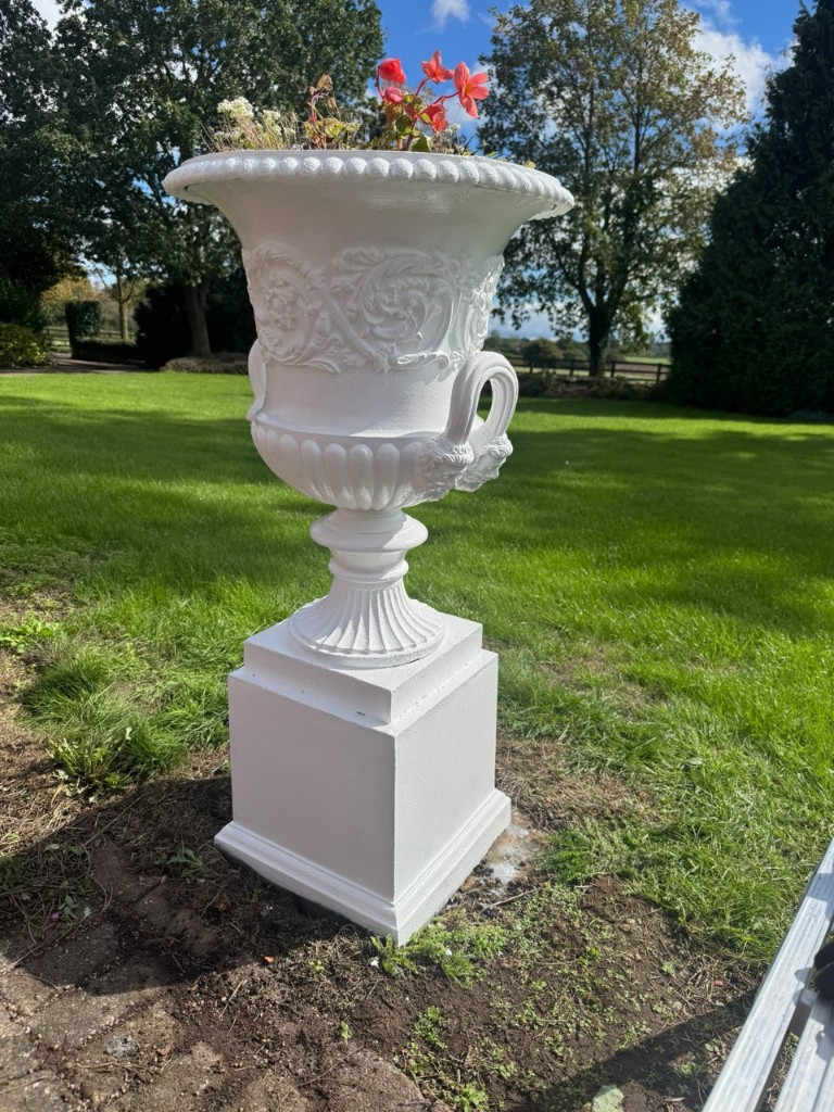 Ornate white-painted cast iron urn planter on a pedestal in a sunny Leicestershire garden